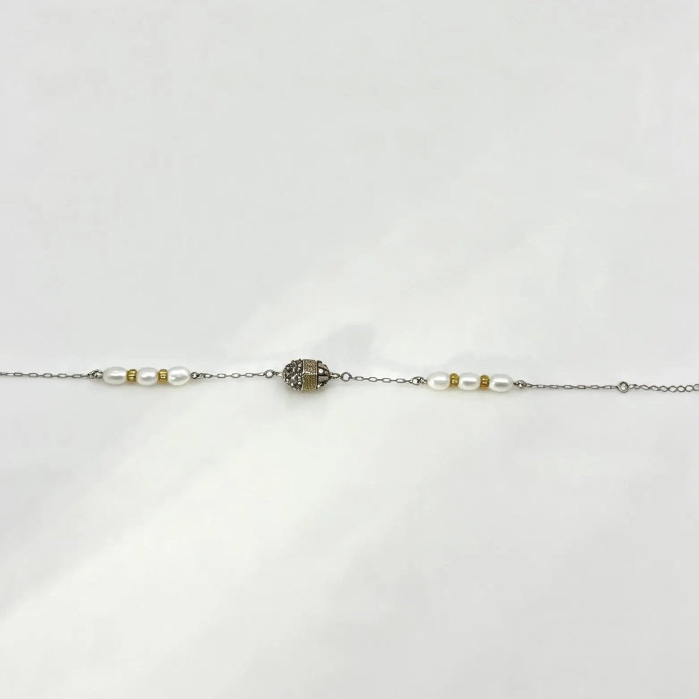 A delicate silver chain bracelet featuring a central oval cluster of small clear stones, flanked by white pearl-like beads and tiny gold spacer beads, laid flat on a white background.